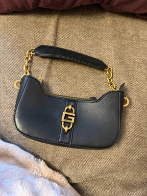 Svart axelväska från Guess - Stilren svart handväska från Guess med guldfärgade detaljer och kedja. Väskan har en elegant form, ett metallspänne med Guess-logga på framsidan och en dragkedja på baksidan. Materialet är slätt och läderliknande, perfekt för att lyfta din outfit.
