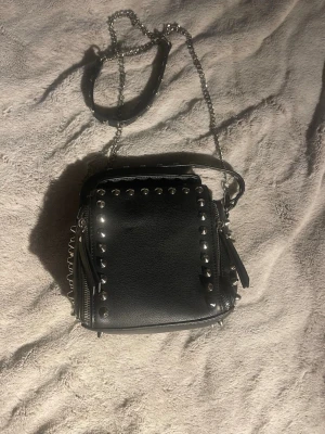 Svart axelväska med nitar och kedja🫶🏼 - Cool svart axelväska i skinnimitation med silverfärgade nitar runt kanterna och en lång kedjerem. Väskan har dragkedjor på sidorna och dekorativa tofsar. Perfekt för dig som gillar en edgy och trendig stil.