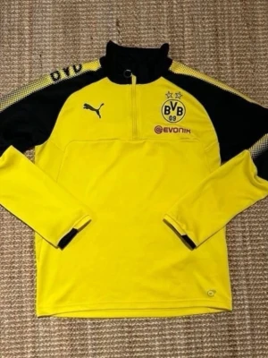 Gul och svart BVB träningströja Puma - Snygg gul och svart långärmad träningströja från Borussia Dortmund (BVB) med halv dragkedja, klubbmärke och Puma-logga på bröstet. Evonik sponsortryck både fram och bak. Tillverkad i funktionsmaterial, perfekt för fotbollsträning.