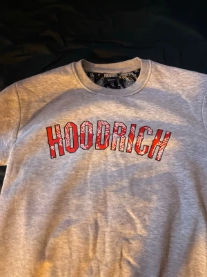 Grå sweatshirt från Hoodrich - Snygg grå sweatshirt från Hoodrich med stort rött och vitt HOODRICH-tryck på bröstet i bandanamönster. Tröjan har rund hals och är tillverkad i mjukt bomullsmaterial. Perfekt för en avslappnad streetwear-look.