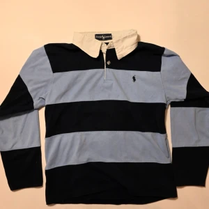 Ralph lauren Randig rugbytröja - Rugbyskjorta från Ralph Lauren med breda marinblå och ljusblå ränder, vit krage och knappar framtill. Klassisk broderad logga på bröstet och långärmad. Tillverkad i bomull för en bekväm känsla.