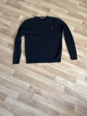Mörkblå stickad tröja från Ralph Lauren - Snygg mörkblå stickad tröja från Ralph Lauren med rund halsringning och diskret röd logga broderad på bröstet. Tröjan har långa ärmar och klassisk kabelstickad struktur som ger en tidlös look. Perfekt att matcha med jeans eller kjol.