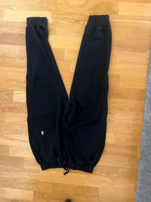 Svarta mjukisbyxor med broderad logga - Säljer ett par svarta mjukisbyxor med resår i midjan och benslut samt dragsko. Byxorna har en liten vit broderad logga på ena benet. Tillverkade i mjukt och bekvämt material, perfekta för chill dagar hemma eller till skolan.