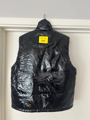 Svart dunväst från Acne Studios - Snygg svart dunväst från Acne Studios med glansig finish och hög krage. Västen har en dragkedja framtill och ett gult smiley-märke på ryggen. Perfekt för lager på lager och streetwear-vibe. Använd en gång bara! Hör av er vid minsta lilla fundering😇