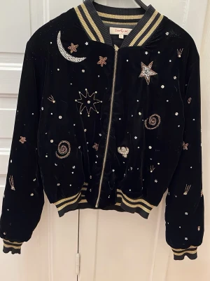 Svart bomberjacka med stjärnbrodyr - Svart bomberjacka i sammet från Daisy Street med broderade stjärnor, månar och galaxmotiv i silver och guld. Jackan har ribbade muddar med guldfärgade ränder och dragkedja framtill. Perfekt statement-plagg för dig som gillar unika detaljer och kosmiska vibbar.