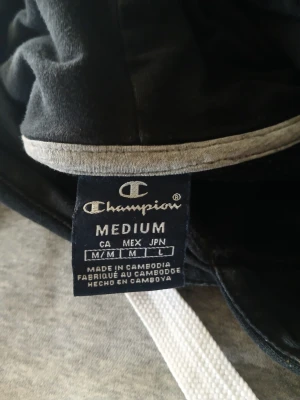 Champion hoodie svart/grå M - Snygg hoodie från Champion i svart och grått med vita detaljer. Stor logga på ena ärmen och mindre logga på bröstet. Klassisk känguruficka framtill och justerbar huva med vita snören. Perfekt för en avslappnad och sportig stil.