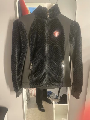 Svart teddyjacka från H Collection - Säljer en svart teddyjacka från H Collection med mjuk, fluffig struktur och detaljer i slätare tyg. Jackan har hög krage, hel dragkedja framtill och två fickor med dragkedja. Märkespatch i brunt läder på bröstet ger en snygg detalj.