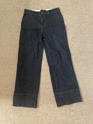 Mörkblå raka jeans med breda slag - Säljer ett par mörkblå jeans med raka ben och breda slag längst ner. Jeansen har hög midja, pressveck framtill och fickor både fram och bak. Klassisk denimlook med kontrastsömmar, perfekta för en trendig och avslappnad stil.
