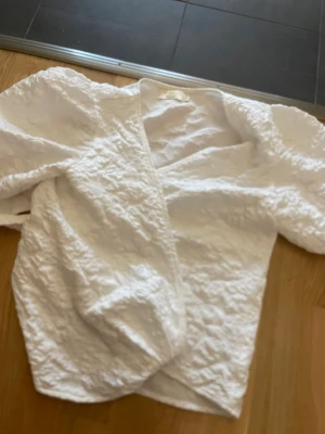 Vit omlottblus med puffärm - Snygg vit omlottblus med puffiga ärmar och struktur i tyget. Blusen har en djup v-ringning och är tillverkad i ett lätt, krinklat material som ger en cool och trendig look. Perfekt att styla med jeans eller kjol för en fräsch outfit.