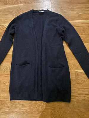 Mörkblå lång kofta från Vila - Mysig mörkblå lång kofta från Vila med öppen front och två fickor framtill. Koftan har ribbade muddar vid ärmslut och nederkant, perfekt att slänga på sig över en t-shirt eller blus. Enkel och stilren design som funkar till det mesta.Den sticks inte 🙌🏻