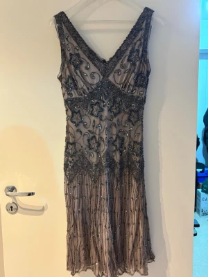 Glittrig kortklänning med pärlor - Superfin kortklänning i ljus beige med glittriga pärlor och paljetter i mörkgrått. Klänningen har v-ringning både fram och bak, är ärmlös och har ett vackert mönster med stjärnor och snirklar. Perfekt för dig som vill sticka ut med en unik look.