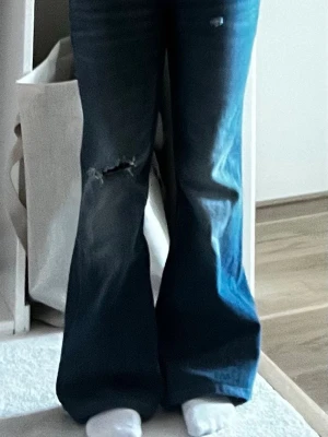 Blå bootcut jeans med slitning - Säljer ett par blå bootcut jeans med slitning och hål på ena knät och ett lite på det andra benet. Jeansen har klassisk femficksdesign och normalhög/ låg midja. 