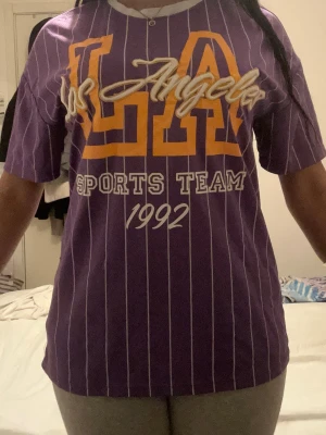 Lila LA Sports Team t-shirt - Säljer en lila t-shirt med vita ränder och tryck framtill med texten 'Los Angeles', 'LA', 'Sports Team' och '1992' i gult och vitt. T-shirten har rund halsringning med vit kant och är kortärmad. Perfekt för dig som gillar sportig streetstyle. Material som även funkar för träning. Storlek S men passar även mig som är M.