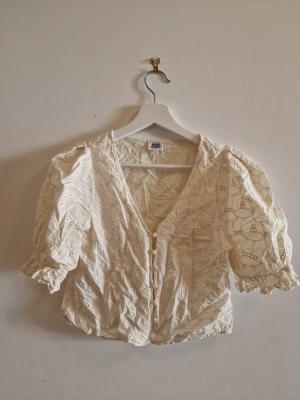 Vit broderad blus från Twist & Tango - Söt vit blus från Twist & Tango med broderade detaljer och v-ringning. Blusen har puffiga trekvartsärmar med resår vid ärmslut och knäppning framtill. Perfekt för en trendig och romantisk look.
