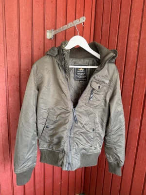Olivgrön bomberjacka Alpha Industries - Snygg olivgrön bomberjacka från Alpha Industries med dragkedja, ribbade muddar och flera fickor. Jackan har en klassisk militärinspirerad look och är tillverkad i slitstarkt material. Perfekt för dig som gillar streetwear och tidlös stil.