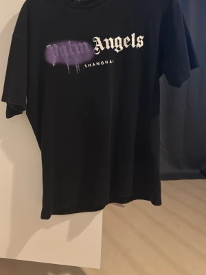 Svart Palm Angels t-shirt Shanghai - Svart t-shirt från Palm Angels med tryck i vitt och lila på bröstet där det står 'Palm Angels Shanghai'. Oversized passform och klassisk rund hals. Perfekt statement-plagg för streetwear-stilen.