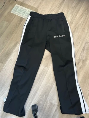Palm Angels svarta mjukisbyxor - Svarta mjukisbyxor från Palm Angels med vita sidoränder och logga tryckt på vänster ben. Byxorna har resår i midjan och raka ben, samt fickor på sidorna. Perfekta för en avslappnad och trendig streetwear-look.
