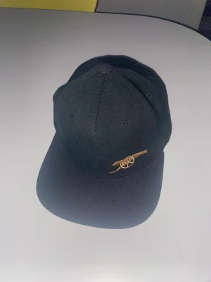 Svart keps från Adidas x Arsenal - Snygg svart keps från Adidas i samarbete med Arsenal. Kepsen har broderad Arsenal-logga i guld framtill och Adidas-logga i guld baktill. Justerbar snapback och klassisk böjd skärm. Perfekt accessoar för dig som gillar sportig stil. Aldrig använd