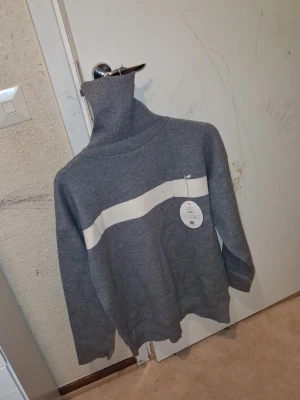 Grå polotröja med vit rand - Snygg grå stickad polotröja med en bred vit rand över bröstet. Tröjan har hög polokrage, långa ärmar och ribbade muddar. Perfekt för dig som gillar en stilren och enkel look. Materialet ser ut att vara mjukt och bekvämt.