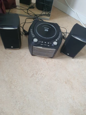 Denver CD-spelare med högtalare - Säljer en svart Denver CD-spelare med två matchande högtalare. Systemet har en rund form på spelaren och rektangulära högtalare. Fronten har digital display, AUX-ingång och flera knappar för enkel styrning. Perfekt för att spela CD-skivor och koppla in andra enheter. OBS! VIKTIGT! cd-spelaren fungerar ej, bara radion. Den här är för någon som vet hur man lagar eller behöver reservdelar. 