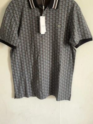 Lacoste Piké - Snygg grå pikétröja från Lacoste med geometriskt mönster i svart och vitt. Tröjan har klassisk krage med vita ränder och korta ärmar. Ikoniska Lacoste-loggan på bröstet. Perfekt för en trendig och avslappnad stil.