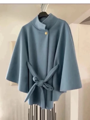 Ljusblå cape-kappa med knytband - Stilren ljusblå cape-kappa med hög krage och ett dekorativt guldknappsfäste vid halsen. Kappan har vida ärmar och ett matchande knytband i midjan för en snygg siluett. Tillverkad i ett mjukt, tjockt tyg som ger en exklusiv känsla.