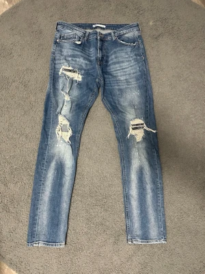 Zara Slitna Jeans M - Säljer ett par blå skinny jeans med slitningar och stora hål på båda benen. Jeansen har klassisk femficksdesign, låg midja och är tillverkade i denim med stretch. Perfekta för en avslappnad och trendig look.