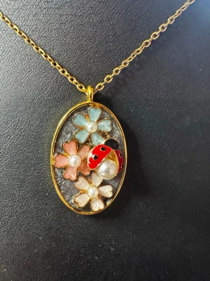 Guldigt halsband med blomma & nyckelpiga - Unikt halsband med en oval guldfärgad ram, dekorerat med pastellfärgade blommor och en söt röd nyckelpiga. Blommorna har pärlliknande detaljer i mitten och hänget har en lekfull, färgglad design. Perfekt accessoar för dig som gillar detaljer och naturinspirerat smycke.