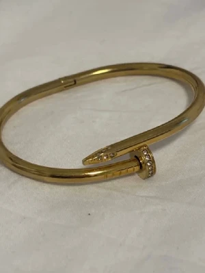 Guldfärgat armband i spikdesign - Snyggt guldfärgat armband i form av en böjd spik, dekorerat med små glittrande stenar på huvudet. 💓 Minsta storleken 