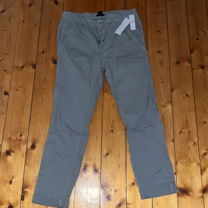 Grå chinos från Lindex, strl 38 - Snygga grå chinos från Lindex i storlek 38. Byxorna har raka ben, klassiska fickor fram och bak samt bälteshällor. Stängs med knappar framtill och är tillverkade i mjuk bomull. Perfekta för en clean och stilren look.