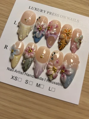 Press-on naglar med blommor och pastell - Snygga press-on naglar i pastellfärger med 3D-blommor, pärlor och bladmotiv. Mix av mandelform och stiletto, handgjorda och återanvändbara. Perfekt för dig som vill ha en unik och kreativ look på naglarna.