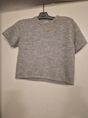 Grå stickad kortärmad tröja - En enkel och stilren grå stickad tröja med korta ärmar och rund hals. Tröjan har en boxig och något croppad passform, perfekt att matcha med jeans eller kjol. Materialet ser ut att vara mjukt och skönt, idealiskt för lager-på-lager. Storlek 146/152