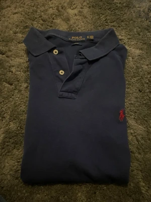 Blå pikétröja från Polo Ralph Lauren - Klassisk blå pikétröja från Polo Ralph Lauren med korta ärmar och krage. Tröjan har två knappar vid halsen och den ikoniska röda logotypen broderad på bröstet. Tillverkad i mjuk bomull som är skön att bära.