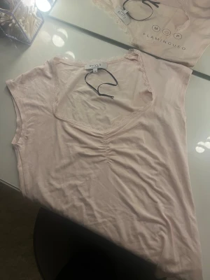 Ljusrosa topp från RYLS med spets - Söt ljusrosa topp från RYLS med v-ringning och spetskant runt halsen. Toppen är ärmlös och har en lätt rynkning framtill som ger en fin detalj. Perfekt att styla med jeans eller kjol för en fräsch look.