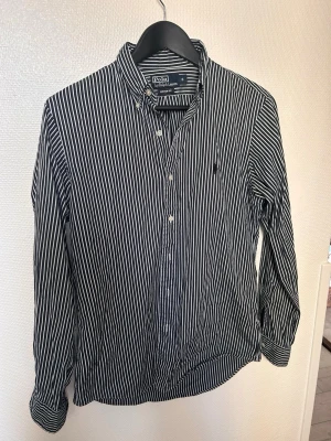 Randig skjorta från Polo Ralph Lauren - Snygg svartvit randig skjorta från Polo Ralph Lauren i modellen Custom Fit. Skjortan har klassisk krage, knäppning framtill och lång ärm. Perfekt för dig som gillar stilrena och tidlösa plagg. Passar även M bra. 