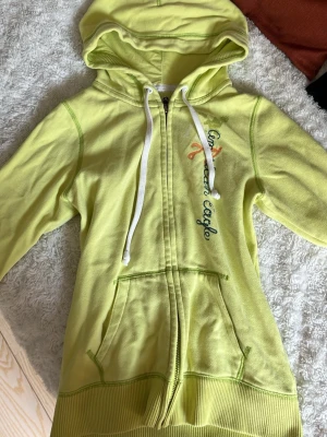 Gul hoodie från American Eagle - Jättefin gul hoodie från American Eagle med broderad logga på bröstet, trekvarts ärmar och vita snören i huvan. Står ej storlek men passar xs/s. Säljer då den inte är i min stil längre. (OBS färgen är mer gul/grön, nästan precis som det syns i bilden men bilden gör så att det syns mer mörk)