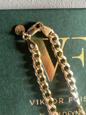 Guldfärgat kedjearmband Viktor Frisk - Stilrent guldfärgat kedjearmband från Viktor Frisk x Guldfynd. Armbandet har platta, breda länkar och en snygg karbinhake. Perfekt accessoar för att lyfta din outfit med en modern touch. Har ny har aldrig använt för den va lite liten för liten för mig.