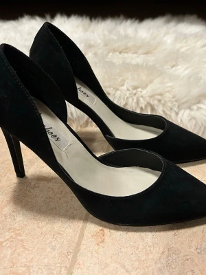 Svarta klackskor, storlek 37 - Svarta klackskor i mockaliknande material och med en elegant öppen design på sidorna, spetsig, stängd-tå, från Nelly. Stilrena och perfekta till fest, middagar och events. Endast testade inomhus, nyskick. Storlek 37.