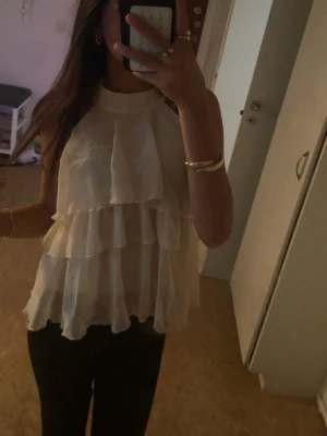 Fest tröja  - Säljer en vit blus med flera lager volanger och hög krage. Blusen är ärmlös och har en luftig, loose passform som ger en cool och trendig look. Perfekt för dig som gillar att sticka ut med detaljer och volanger.