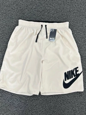 Beiga Nike shorts med logga - Snygga beiga shorts från Nike med stor svart logga på vänster ben. De har elastisk midja med svarta dragsnören och är tillverkade i ett lätt och bekvämt material, perfekt för en avslappnad och sportig look.