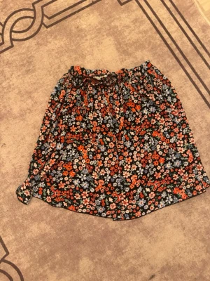 Blommig kortkjol från ONLY - Supersöt kortkjol från ONLY med blommigt mönster i orange, blått och vitt på svart botten. Kjolen har resår i midjan och är perfekt för dig som gillar färg och mönster. Lätt och luftig modell som passar till många olika stilar.