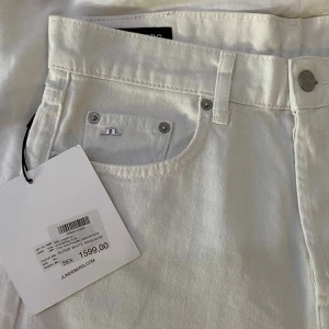 Vita jeans från J.Lindeberg - Snygga vita jeans från J.Lindeberg i modellen Cloud White. Klassisk femficksdesign med silverfärgade detaljer och läderpatch bak i midjan. Byxorna har bälteshällor och är tillverkade i bomull för en skön känsla. Perfekta för en clean och fräsch look. Prislappen är inte kvar och jeansen är använda en gång utan några skillnader på utseenden.