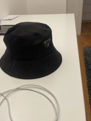 Svart bucket hat från Prada - Snygg svart bucket hat från Prada med klassisk siluett och diskret logga framtill. Hatten är tillverkad i ett slätt tyg som ger en stilren look och passar perfekt till streetwear eller trendiga outfits.