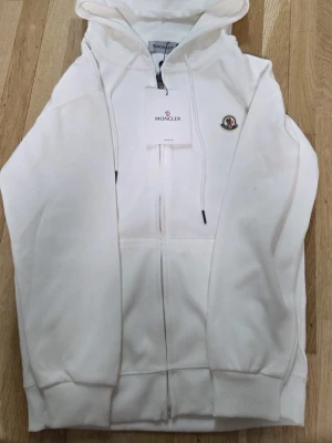 Vit hoodie med dragkedja - Snygg vit hoodie, med dragkedja framtill och klassisk logga på bröstet. Modellen har huva med dragsko och ribbade muddar vid ärmslut och nederkant. Perfekt för en clean och stilren look.