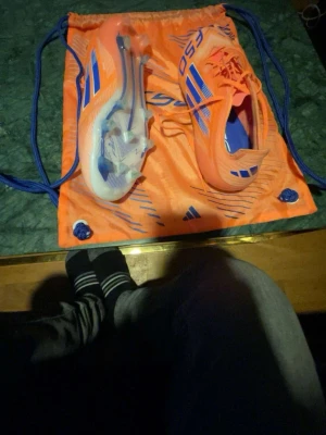 Adidas X Speedflow orange fotbollsskor - Säljer ett par Adidas X Speedflow fotbollsskor i en riktigt snygg orange färg med blå detaljer och vita sulor. Skorna har snörning och är tillverkade i ett lätt syntetmaterial för maxad speed på planen. Perfekta för dig som vill sticka ut och spela snabbt!