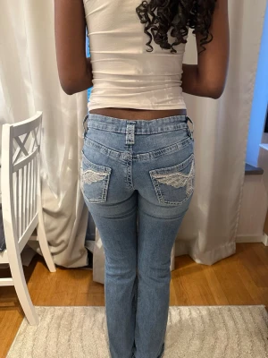 Ljusblå bootcut jeans med brodyr - Säljer ett par ljusblå bootcut jeans med slitningar på knäna och snygg vit brodyr på bakfickorna. Jeansen har låg midja och klassisk femficksdesign. Perfekta för en avslappnad och trendig look. Använda ett par gånger och är storleken 170 på Young Gina.
