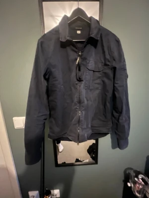 Mörkblå vindjacka från C.P. Company - Snygg mörkblå vindjacka från C.P. Company med dragkedja framtill, bröstficka och klassisk linsdetalj på ärmen. Jackan har en stilren design och är tillverkad i lätt, vindtätt material som passar perfekt för blåsiga dagar.
