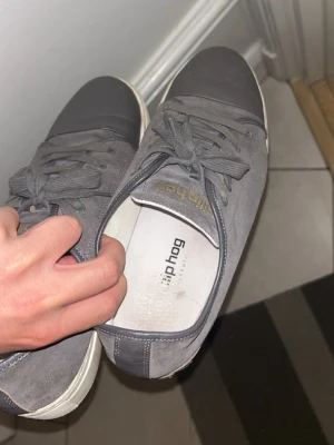 Grå sneakers från Filip Hag - Snygga grå sneakers från Filip Hag med ovandel i mocka och tåparti i slätt material. Klassisk låg modell med vita sulor och matchande grå skosnören. Diskret guldfärgad logga på sidan. Perfekta för en clean och stilren look.