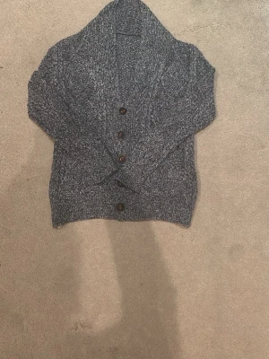Mörkgrå stickad kofta med knappar - Mysig mörkgrå stickad kofta med grov struktur och stora bruna knappar framtill. Koftan har en bred krage och två fickor framtill. Perfekt för lager på lager och chill stil. Passar från 168 till 175