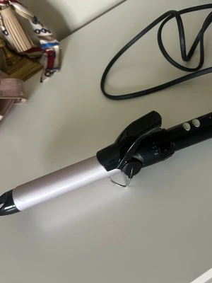 Locktång i svart och silver - Säljer en locktång med svart handtag och silverfärgad värmecylinder. Den har en klämma, två knappar för inställning och en lång sladd. Perfekt för att fixa snygga lockar snabbt och enkelt.